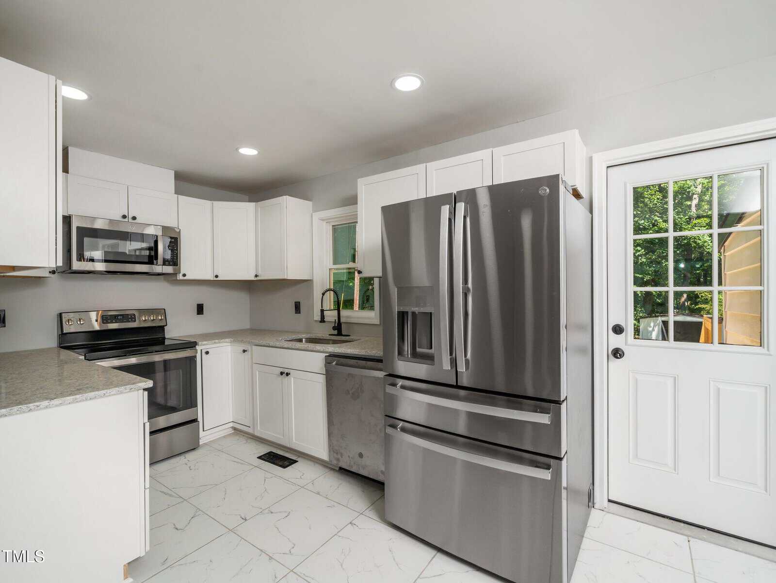 Modern kitchen with white cabinets, stainless steel appliances, gray countertops, a large refrigerator, and a dishwasher. There is a window above the sink and a glass door leading outside. The floor is white with gray veining.