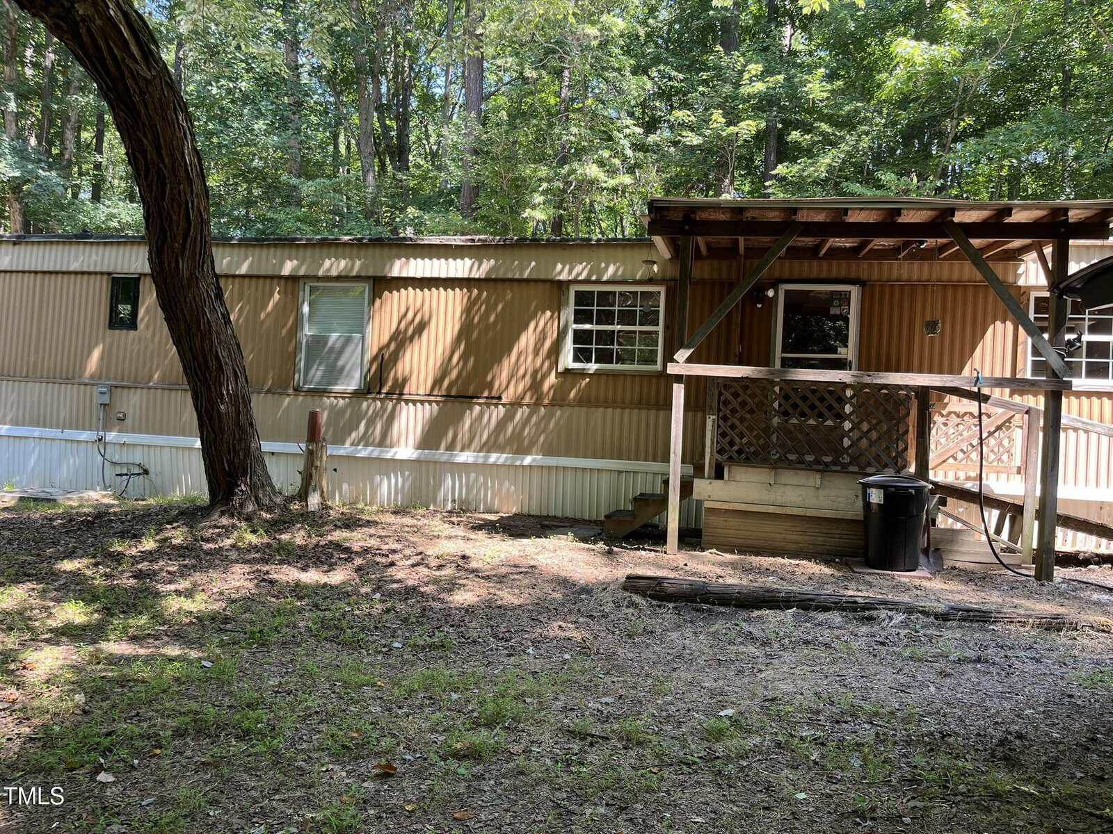 Single-wide tan mobile home with a covered wooden porch, set in a wooded area with tall trees and dappled sunlight. A large tree is in the foreground, and there is a trash can near the steps.