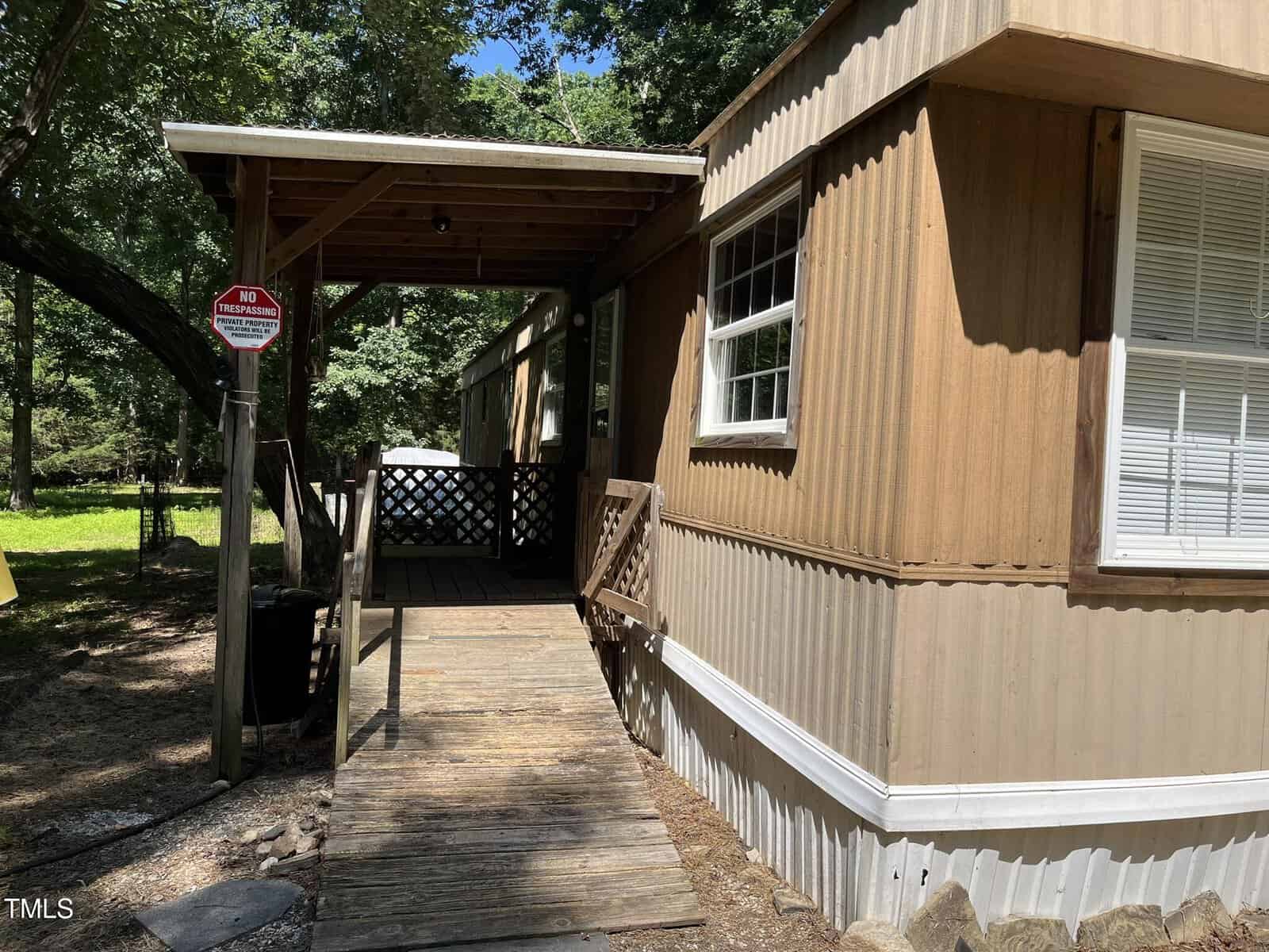 A tan mobile home with a wooden ramp leading to a covered porch. A "No Trespassing" sign is posted on a pole near the ramp. Trees and a grassy yard are visible in the background.