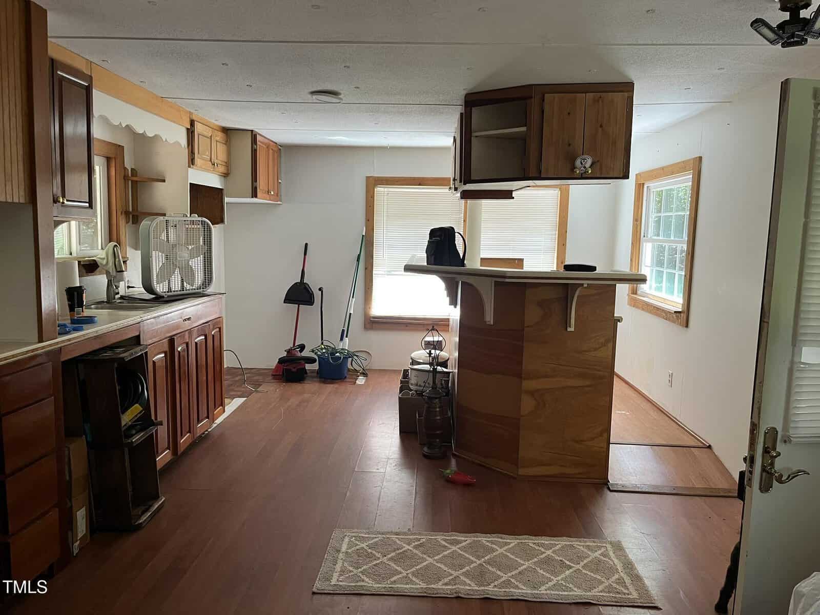 A small, unfurnished kitchen and dining area with wood cabinets, a built-in bar, cleaning supplies against the back wall, a fan on the counter, and light streaming through two large windows.