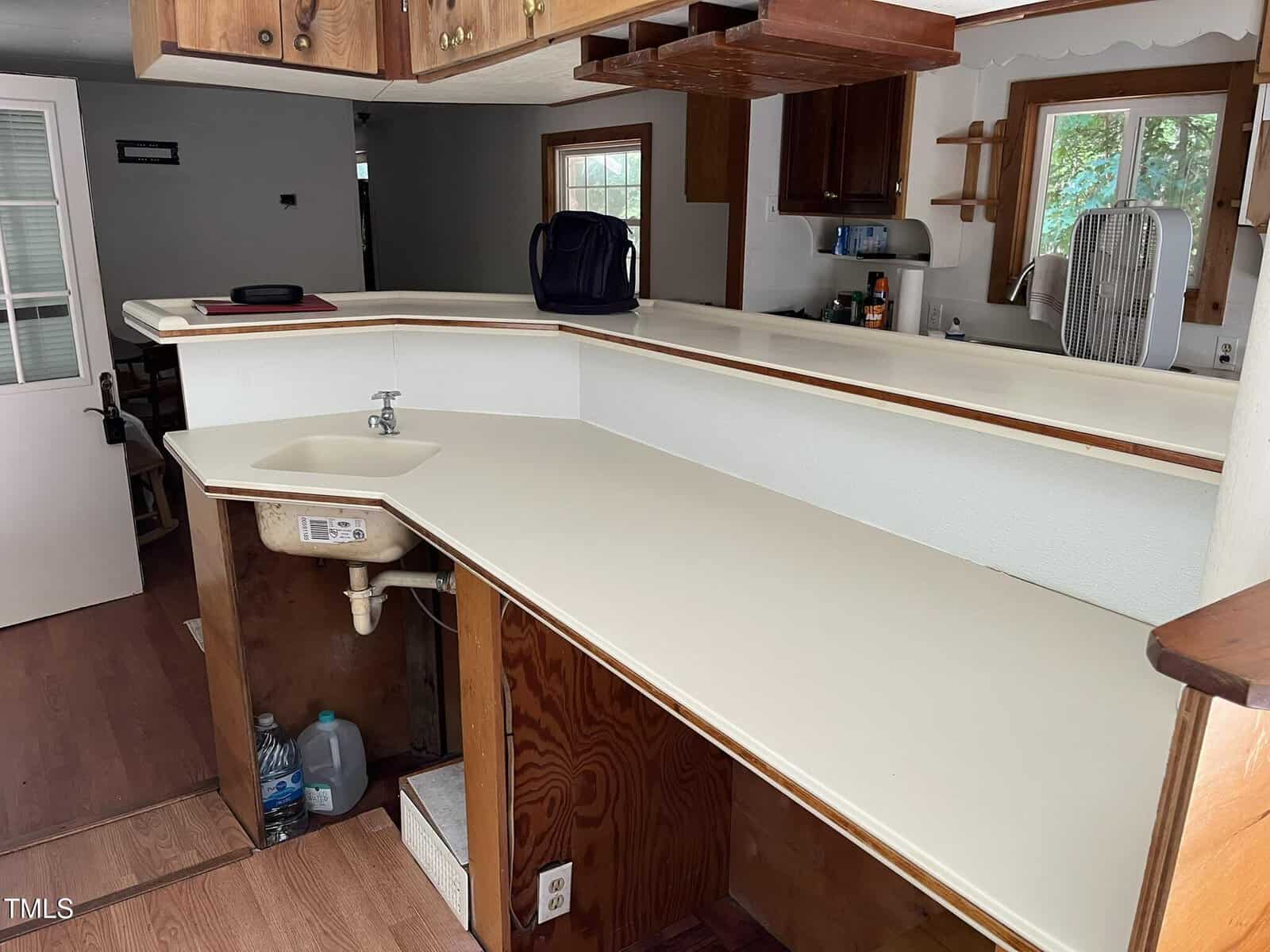 A kitchen with a white countertop, a sink, and open cabinets underneath. There is a raised bar, some wooden cabinets above, a bag on the counter, and a window with trees visible outside.