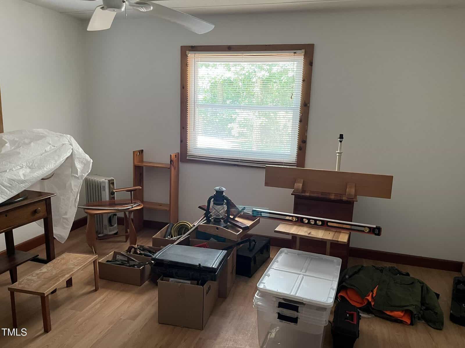 A sparsely furnished room with a window, a ceiling fan, and wooden trim contains scattered boxes, tools, a lamp, a heater, a covered item, a coat, and a few small pieces of furniture on a light wood floor.