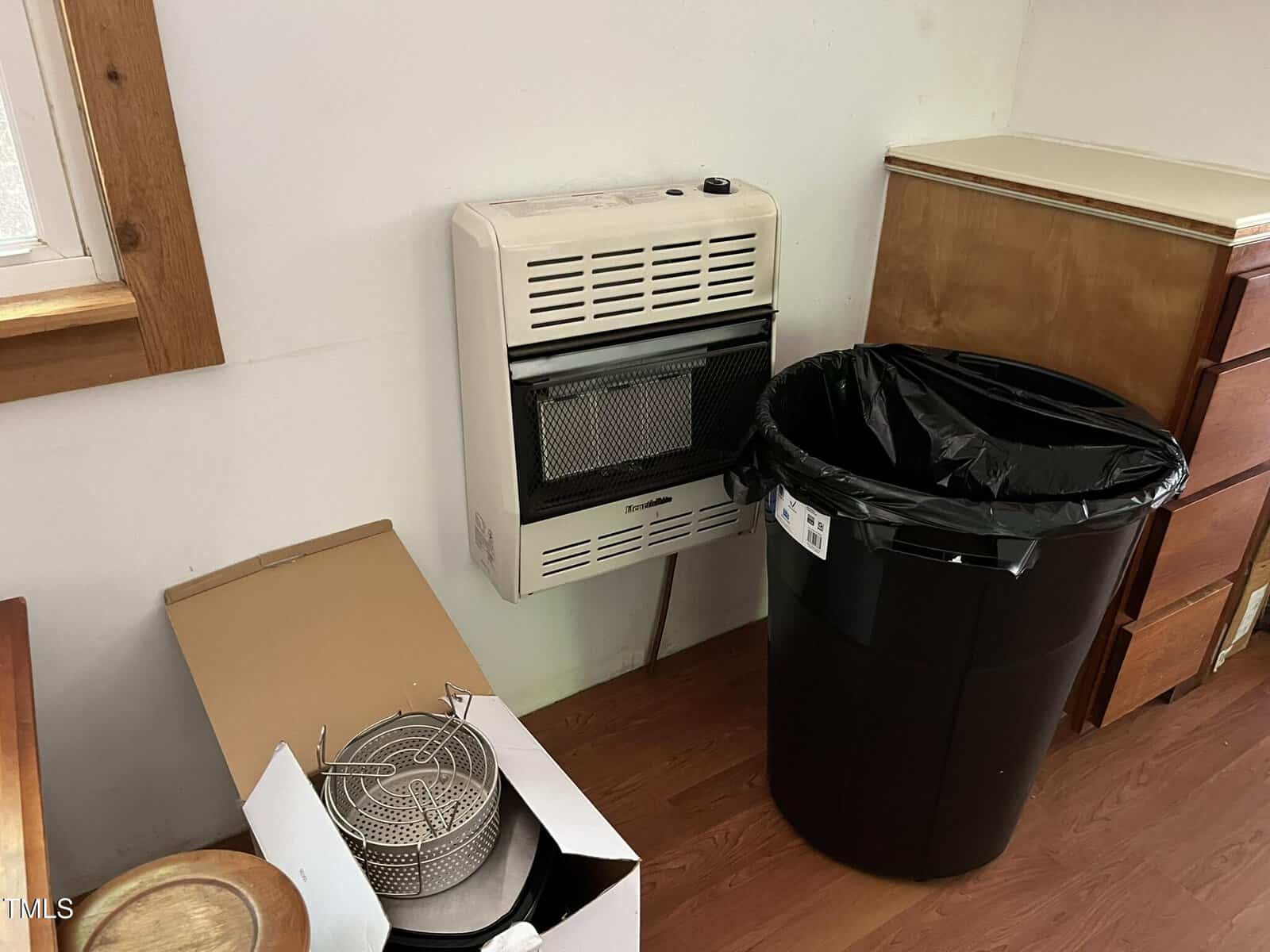 A small wall-mounted heater next to a large black trash can with a black liner, a wooden dresser, and an open box containing metal kitchen items, all on a wood floor in a corner of a room.