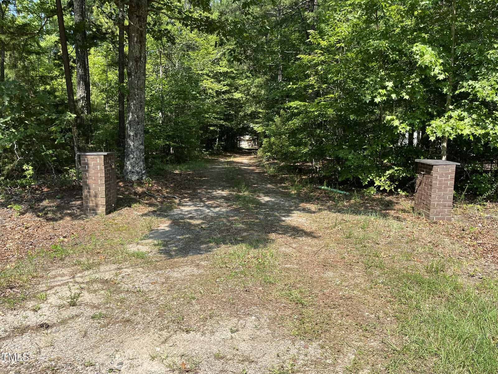 A gravel driveway leads into a wooded area, flanked by two brick pillars on either side, with dense green trees and foliage surrounding the entrance. Sunlight filters through the leaves, creating dappled shadows on the path.
