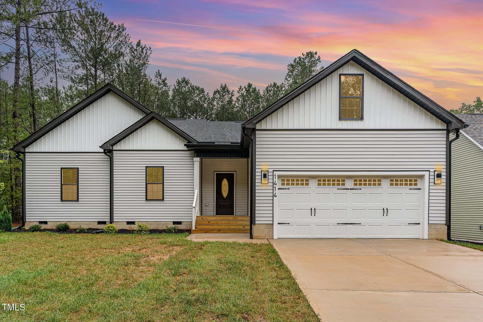 Single-story modern house at 1414 Sagamore Drive, Louisburg, with white siding, black trim, a double garage, and small front porch. The yard features a concrete driveway, grass, trees, and a colorful sky in the background.