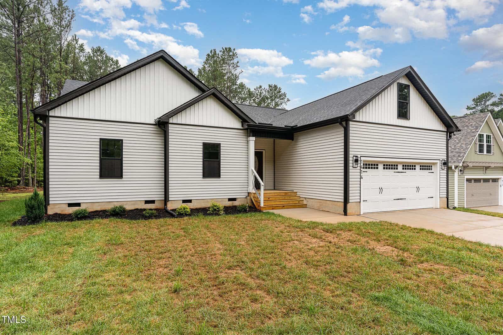 Single-story house with white vertical siding, black trim, and a two-car garage. The home has a small covered front entry, steps leading to the door, and a driveway, with grass and shrubs in the front yard.