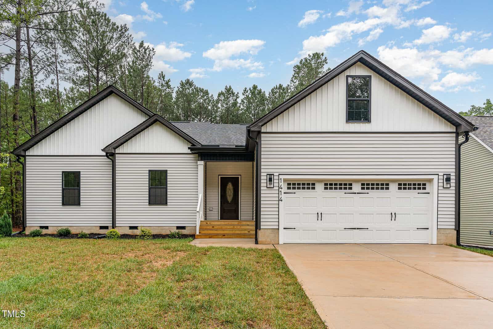 A single-story white house with black trim, a front porch, a two-car garage, concrete driveway, and a grassy yard, set against a backdrop of tall trees and a partly cloudy sky.
