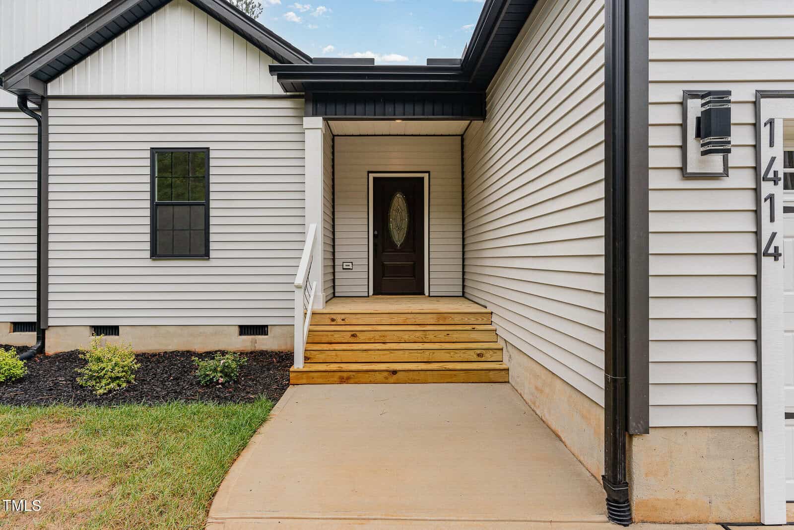 A concrete walkway leads to a wooden front porch with steps and a dark-colored front door. The house has light-colored siding, black trim, a window, and a house number sign reading 1414. Shrubs are planted along the side.