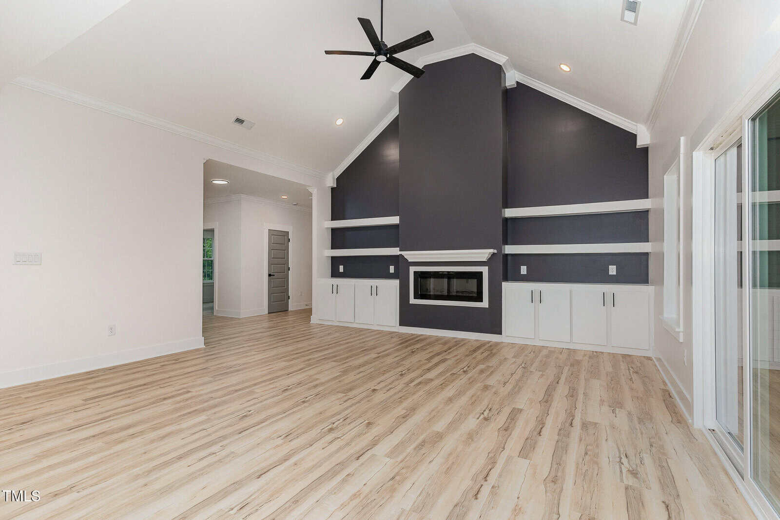 Spacious modern living room with light wood floors, a vaulted ceiling, a black ceiling fan, and a dark accent wall featuring a built-in fireplace, shelves, and white cabinetry. Large windows and doors line the right wall.