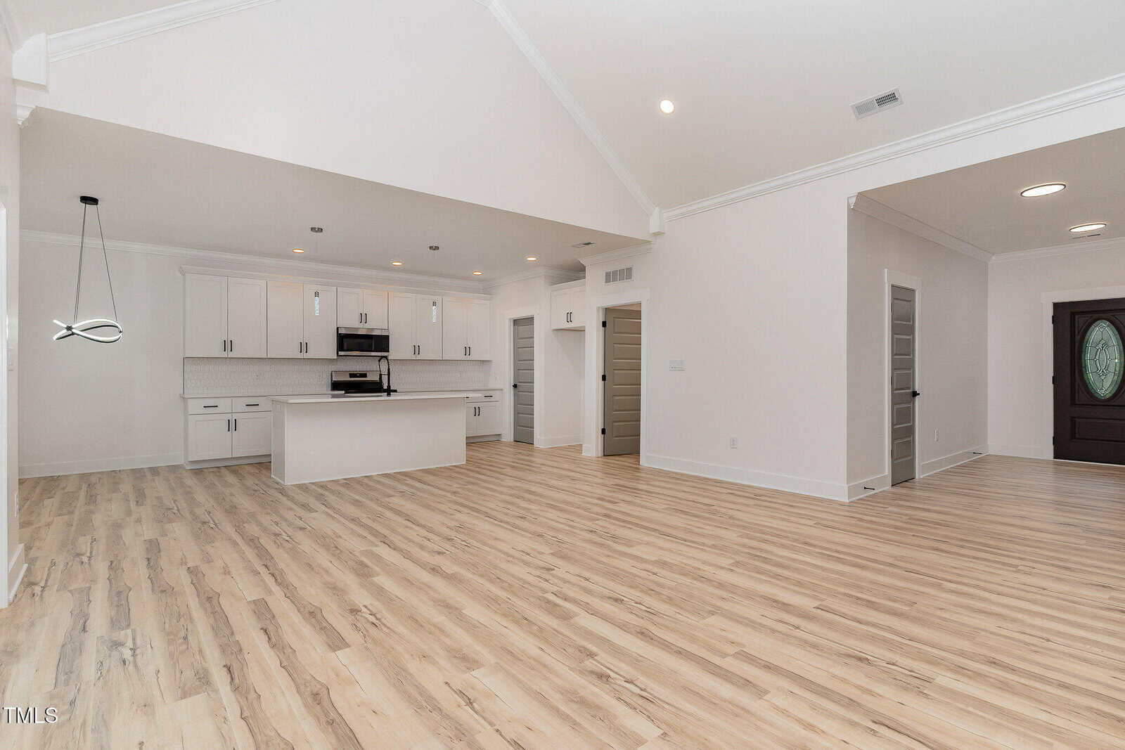 Spacious open-concept kitchen and living area with light wood floors, white walls, recessed lighting, and a central island. Modern cabinets and stainless appliances are visible; multiple doors lead to other rooms.