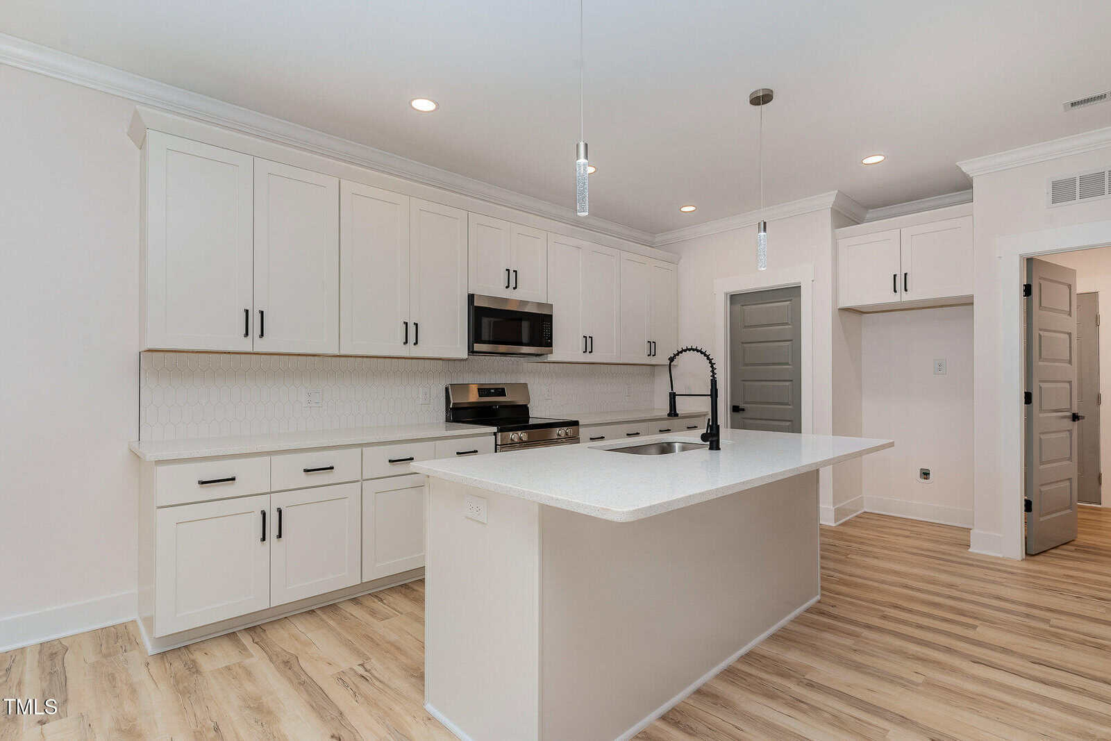 A modern kitchen with white cabinets, stainless steel appliances, a white island with a black faucet, light wood flooring, and two pendant lights hanging from the ceiling. Multiple gray doors are visible in the background.