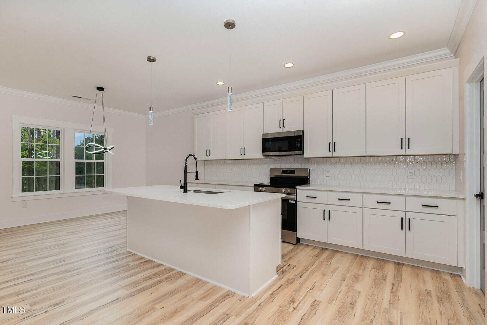 Modern kitchen with white cabinets, black hardware, stainless steel appliances, a large island with sink and black faucet, light wood flooring, white tile backsplash, and pendant lights above the island.