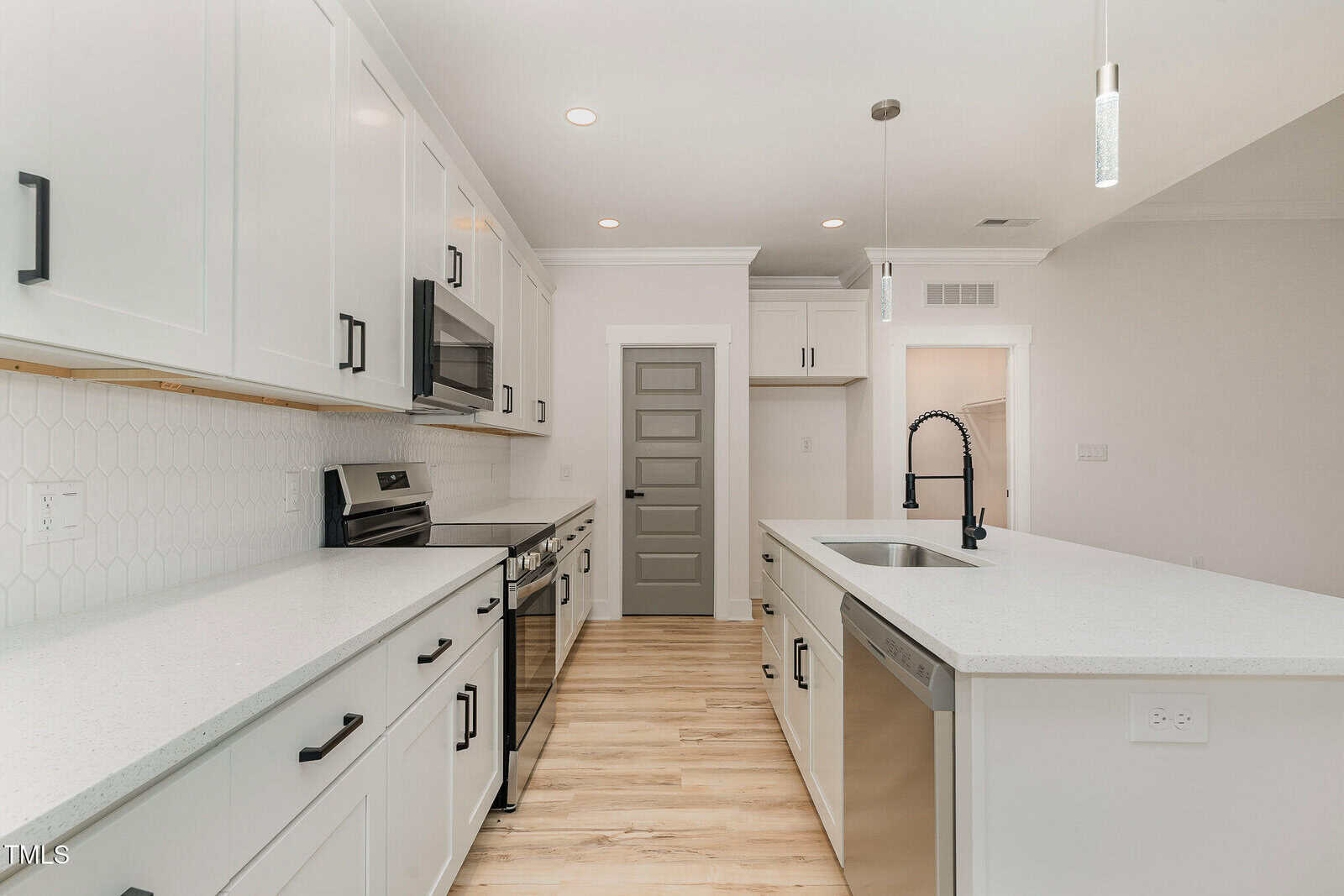 Modern kitchen with white cabinets, stainless steel appliances, island with sink and dishwasher, light wood flooring, and hexagonal tile backsplash. Gray door at the end of the room. Bright and spacious layout.
