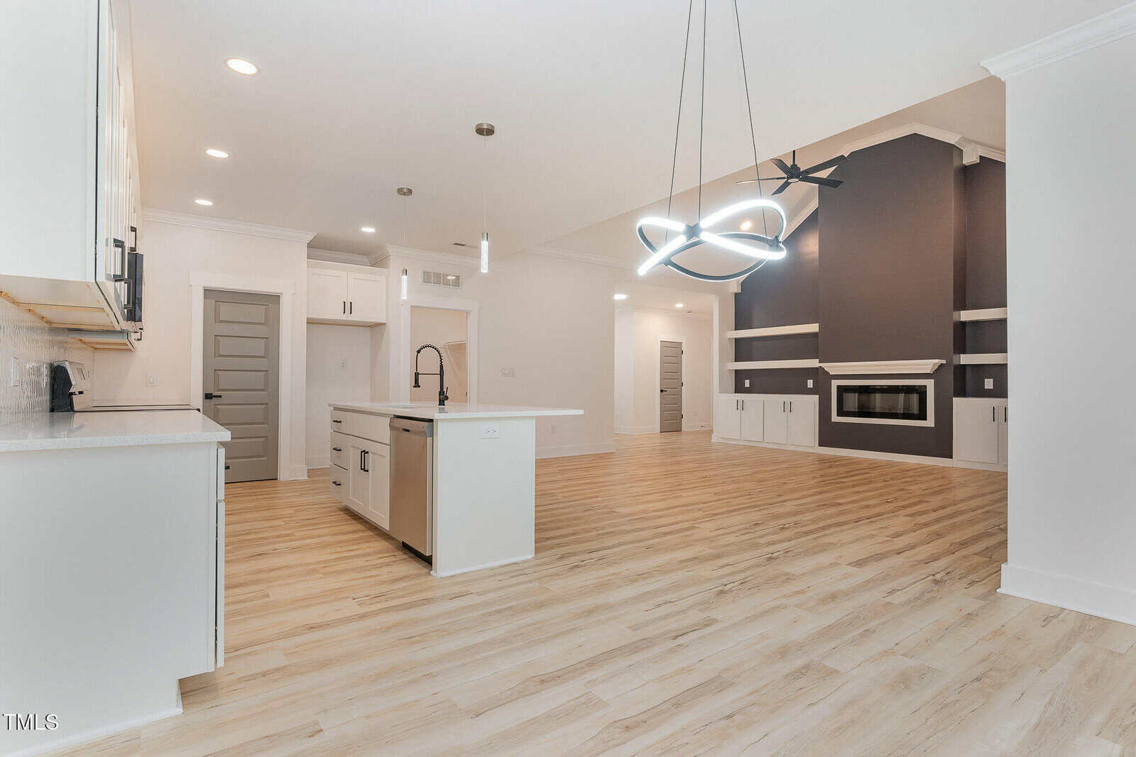 Modern open-concept kitchen and living area with light wood flooring, white cabinets, a central island, sleek lighting fixtures, and a dark accent wall with a built-in fireplace and shelves.