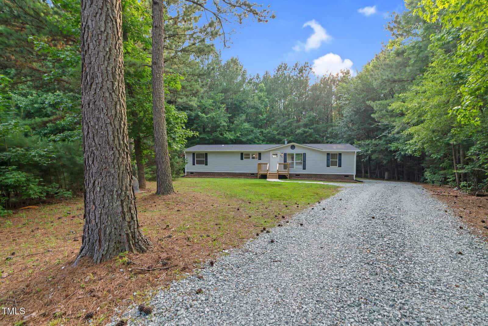 A single-story light gray house with a front deck sits next to a gravel driveway surrounded by tall trees and dense forest under a blue sky with some clouds.