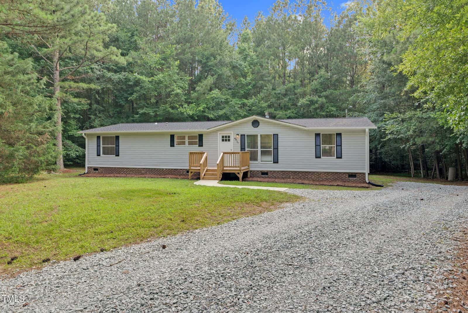 A single-story house with light gray siding and dark shutters sits in front of a wooded area. A small wooden porch with steps leads to the front door, and a gravel driveway curves toward the house.