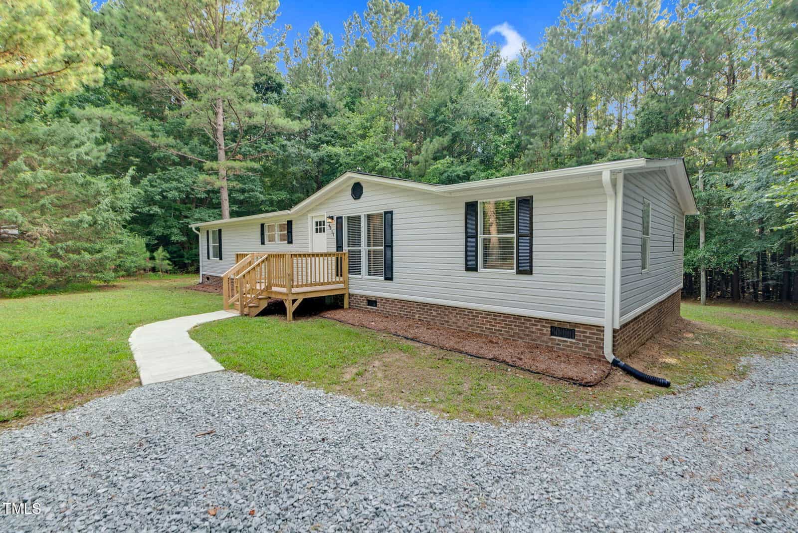 A single-story house with white siding and a brick foundation is surrounded by trees. The house has a small wooden porch with steps, large windows, and a gravel driveway leading to a concrete walkway.