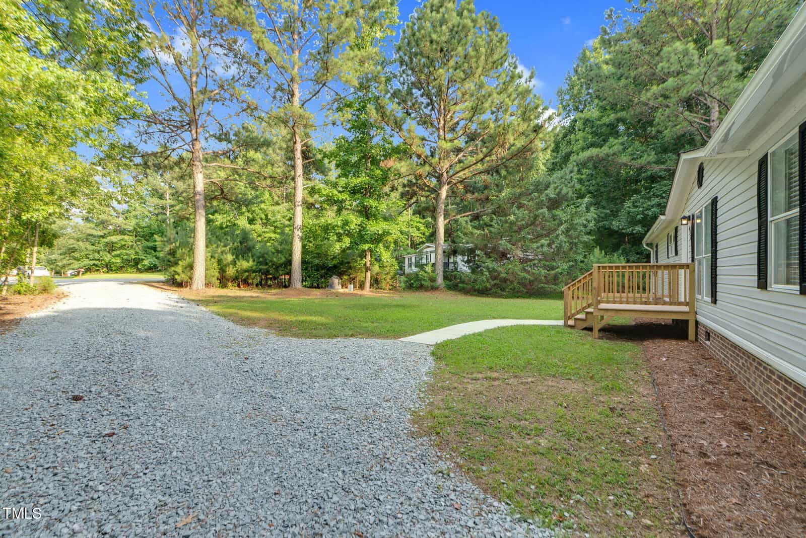 A gravel driveway curves past a white single-story house with a small wooden deck. Tall pine trees and dense greenery surround the area under a clear blue sky.