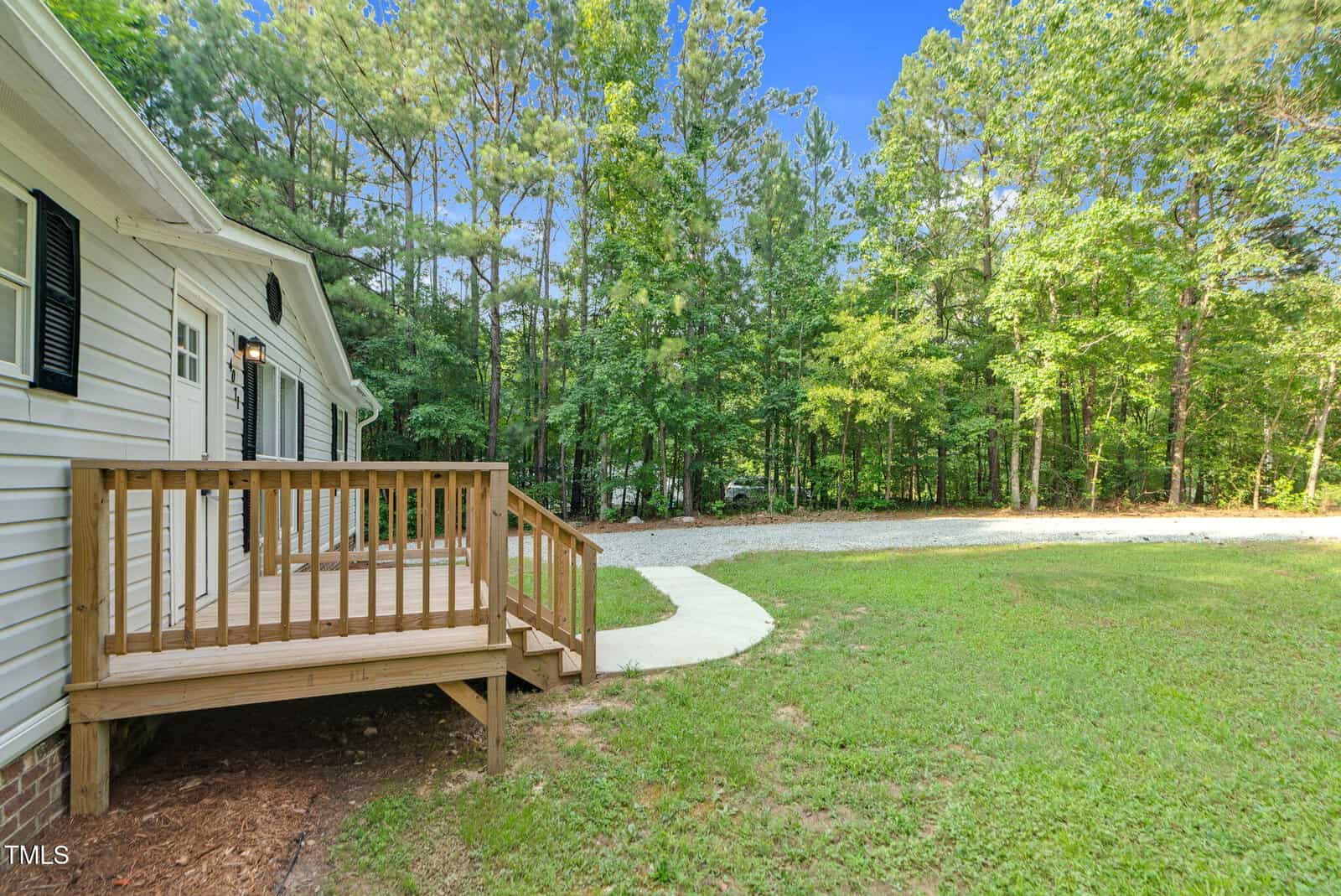 A small wooden porch with steps leads to a white-sided house, facing a curved concrete walkway and a gravel driveway. The yard is grassy, bordered by dense green trees under a clear blue sky.