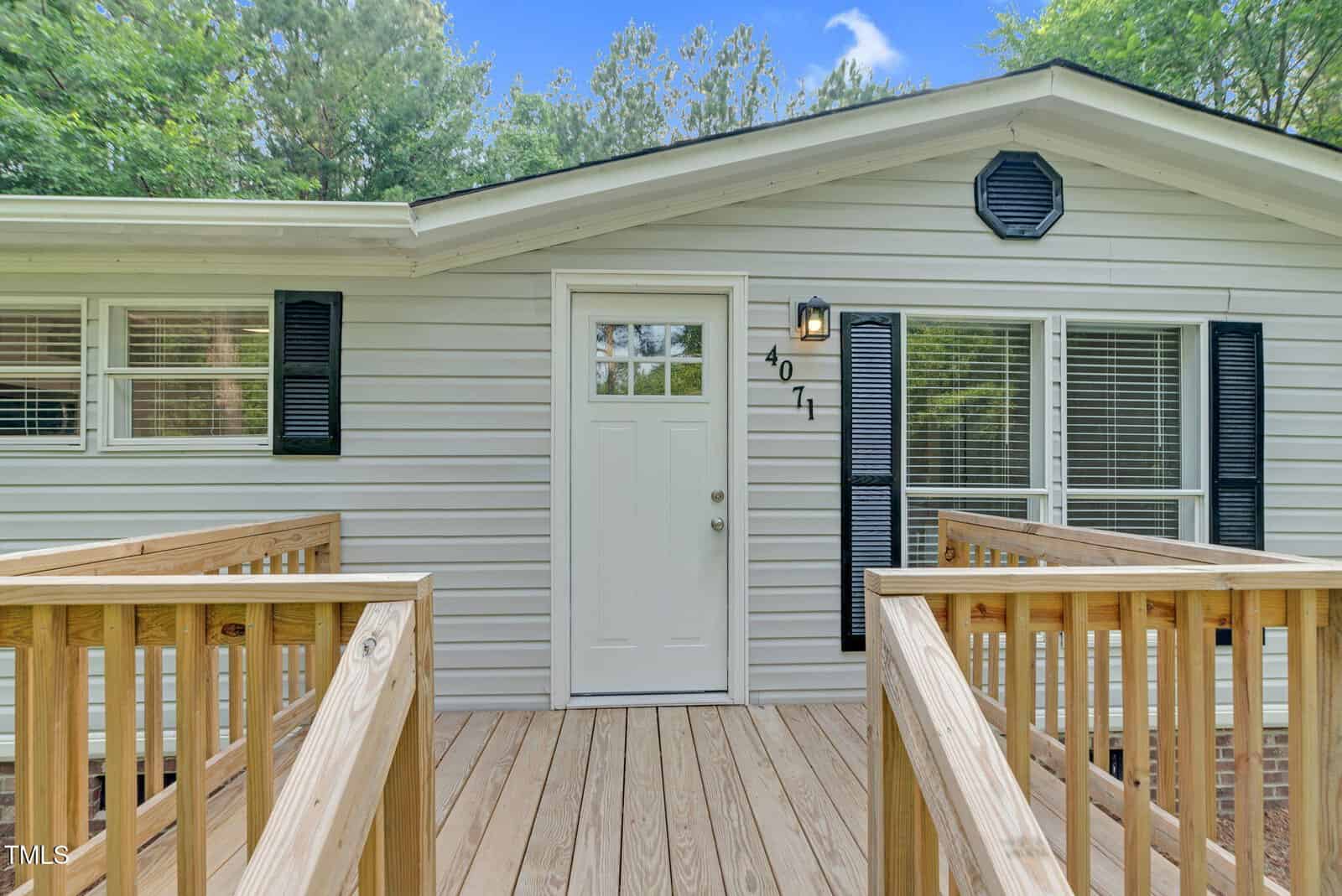 A white house with black shutters and a central white door is shown. A wooden ramp leads up to the entrance. The number 4011 is next to the door. There are trees in the background and a blue sky with clouds.