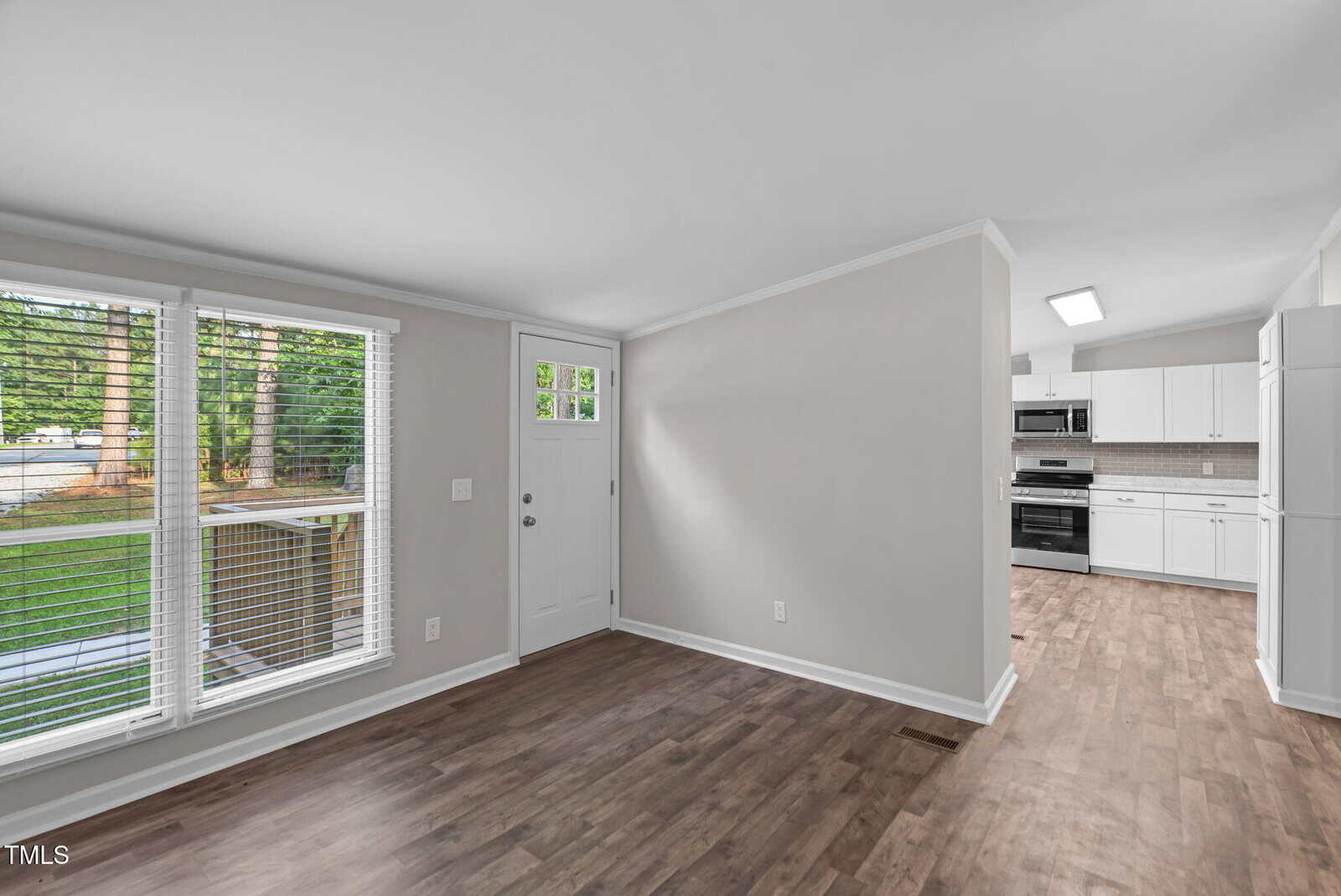 A view of a room with light gray walls, wood flooring, large windows with white blinds, a white door, and an open entryway leading to a kitchen with white cabinets and stainless steel appliances.