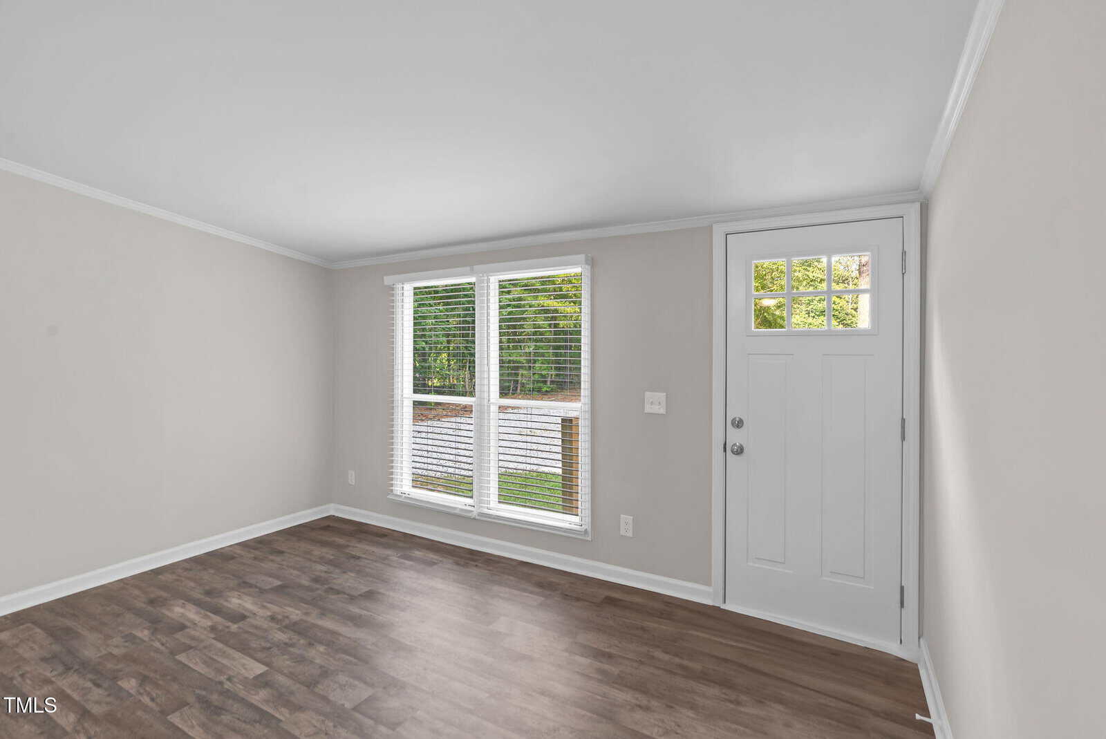A small, empty room with light gray walls, wood-style flooring, two large windows with blinds, and a white door with a window panel. Natural light comes in through the windows.