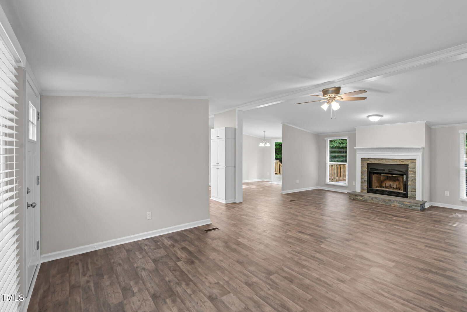Spacious living area with wood floors, neutral walls, ceiling fan, and a fireplace. Large windows provide natural light, and open doorways lead to kitchen and dining spaces.