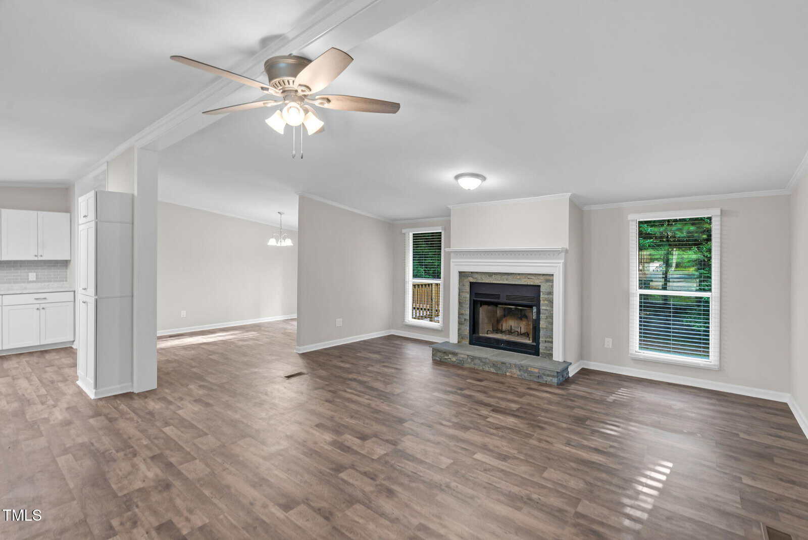 Bright living room with wood floors, ceiling fan, fireplace, and two large windows. Open layout leads to a modern white kitchen and dining area. Neutral walls and natural light create a spacious, inviting feel.