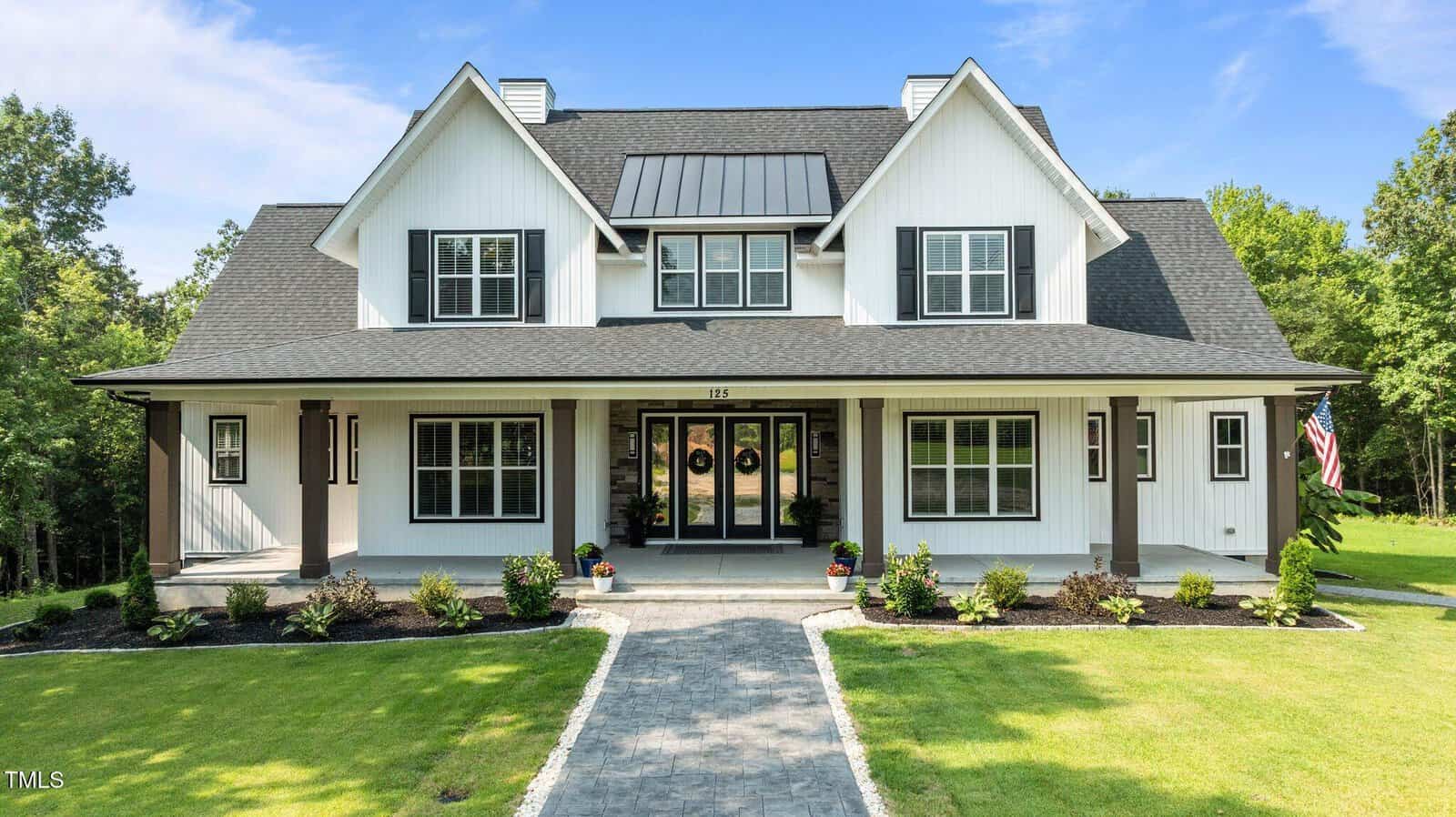 A modern white two-story house with black accents, a covered front porch, large windows, and a landscaped yard with green grass, shrubs, and a stone walkway leading to the entrance. An American flag is visible on the right.
