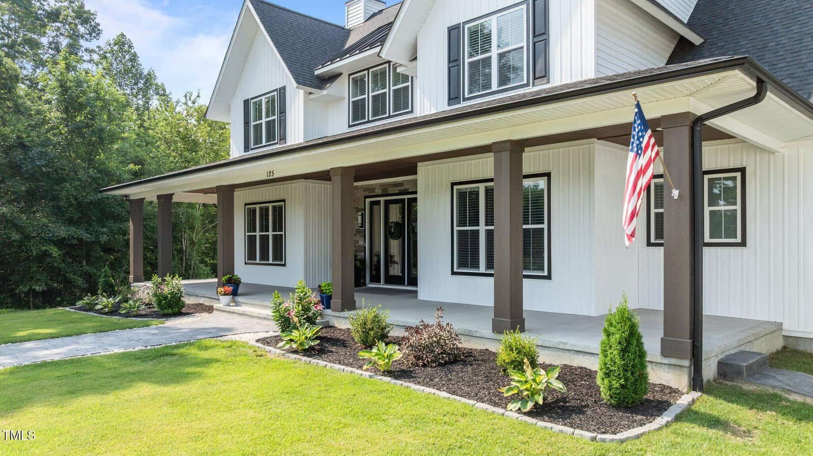 A modern white two-story house with black trim, a covered front porch supported by dark columns, a manicured lawn, landscaping, and an American flag by the entrance. Trees are visible in the background.