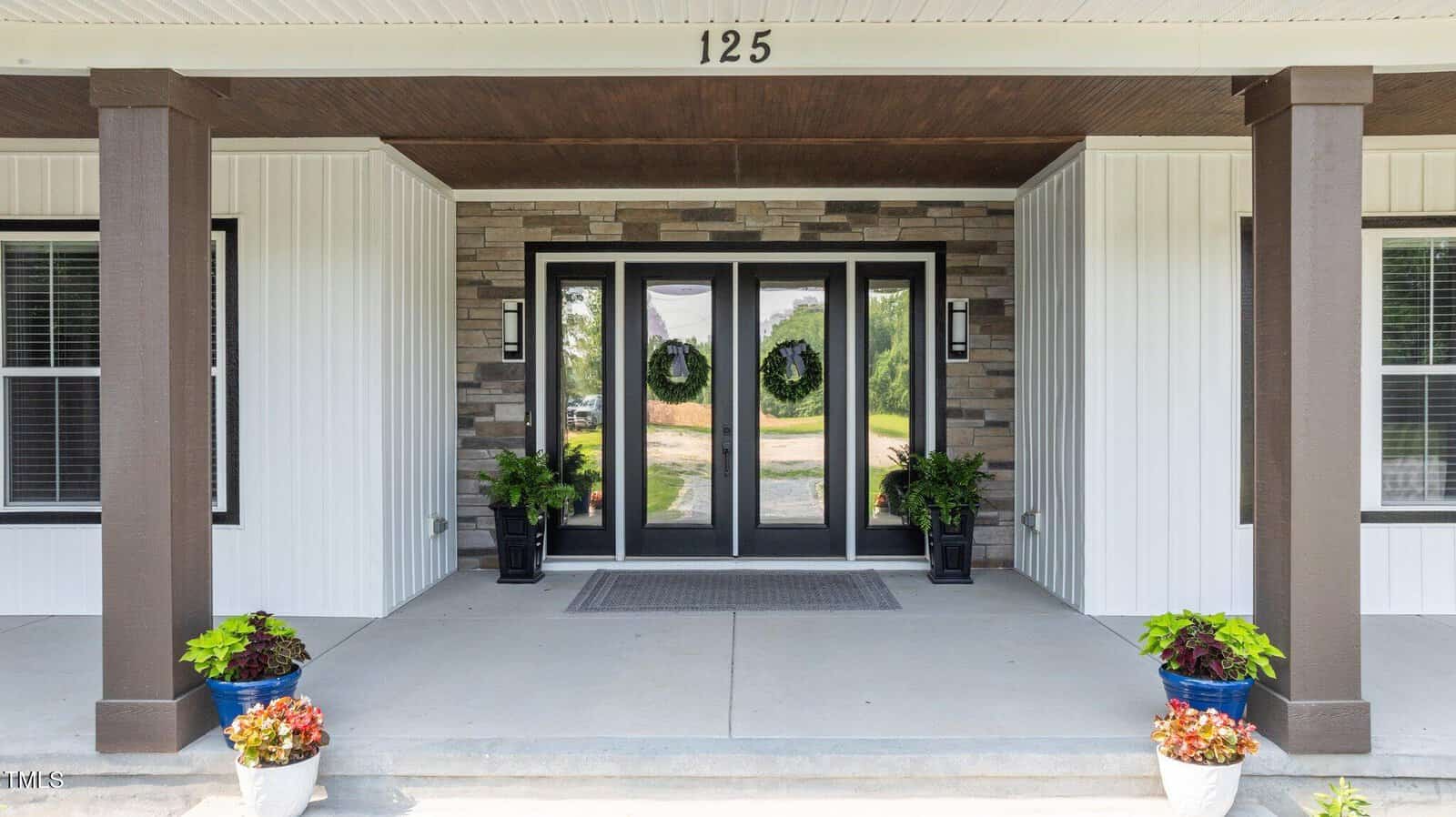 Front porch of a house with stone and white siding, double glass doors with wreaths, two windows, potted plants and flowers on either side of the entrance, and the number 125 above the door.