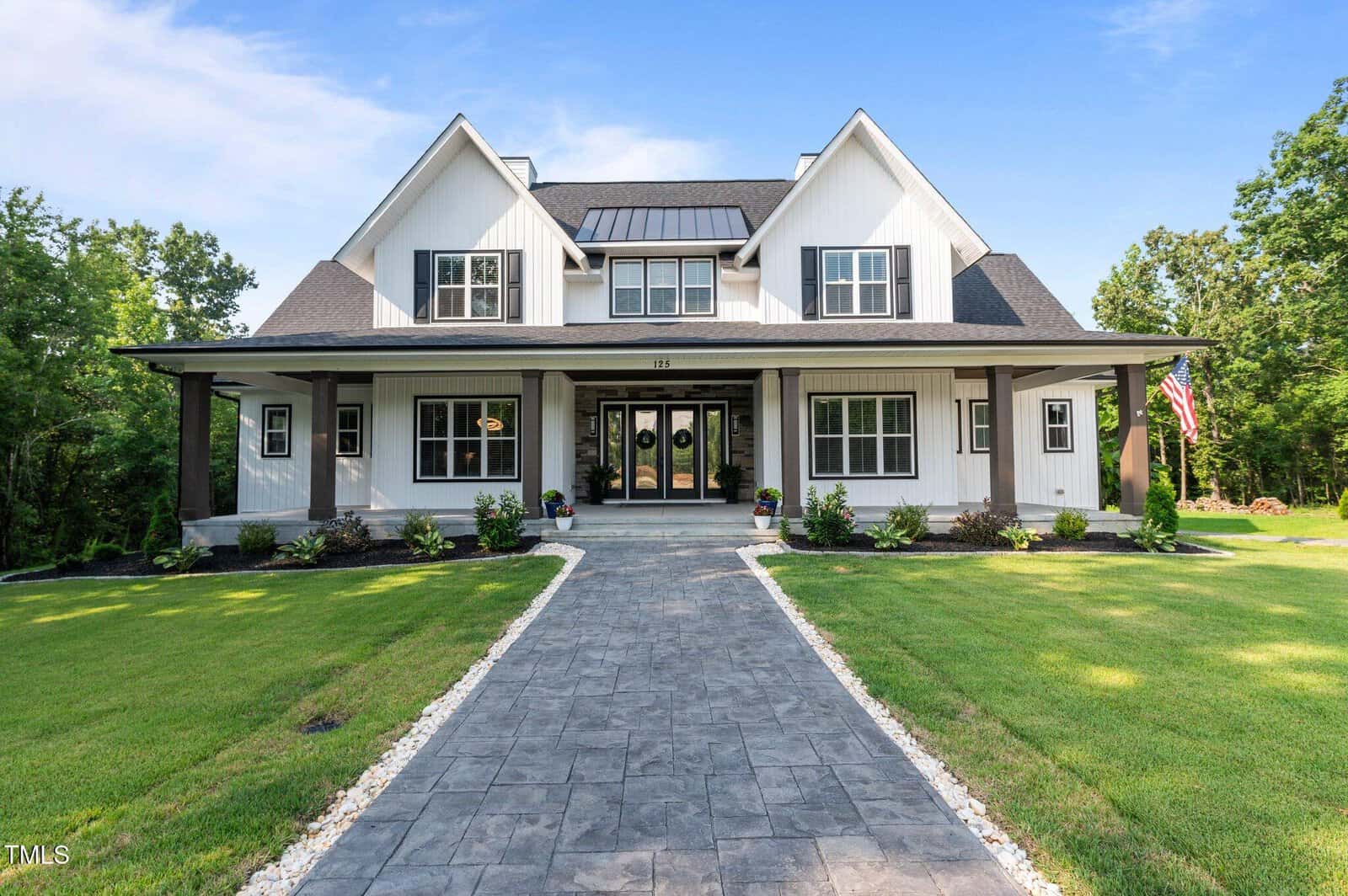 A modern two-story farmhouse with white siding, dark trim, a large covered front porch, double front doors, neat landscaping, a stone walkway, and an American flag on the right side, set against a green lawn and trees.