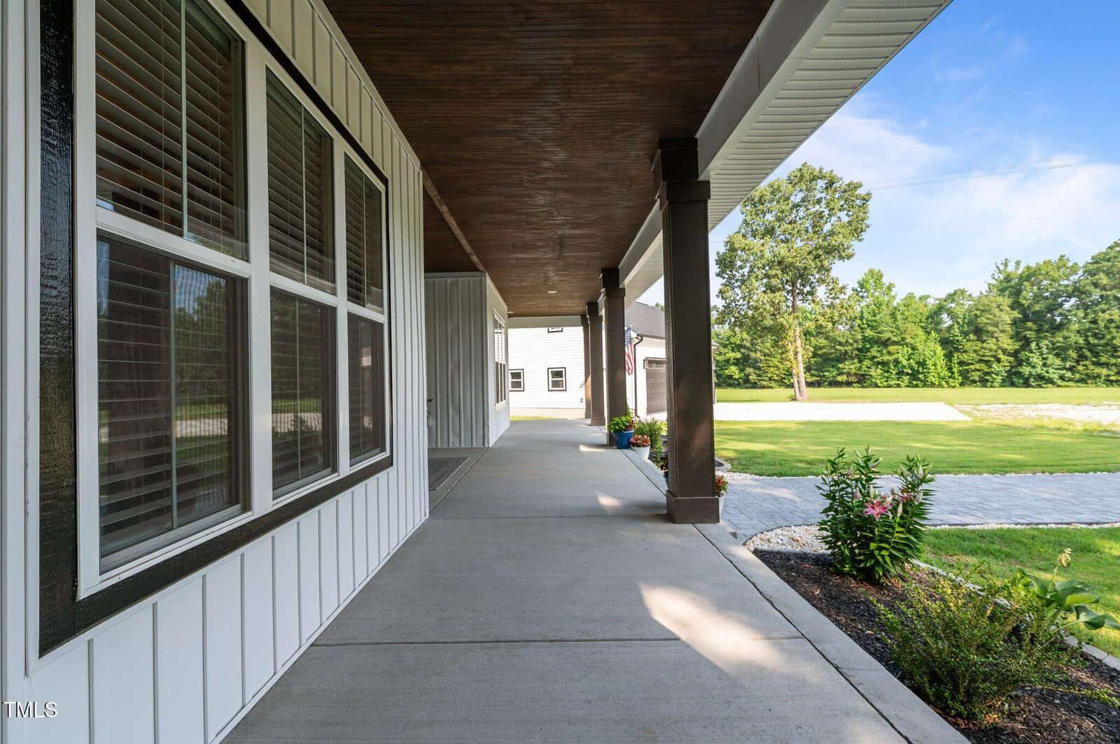 Wide view of a modern house's covered front porch with white siding, large windows, wood accents, potted plants, and a landscaped yard with a stone path and green lawn under a clear sky.