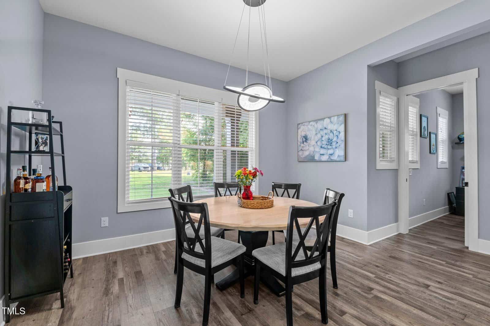 A dining room with a round wooden table, six black chairs, a modern pendant light, a bar cart in the corner, and a large window with white blinds, letting in natural light. Walls are light gray with simple decor.