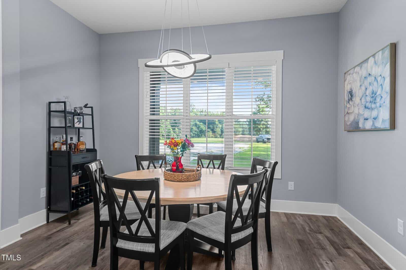 A dining room with a round wooden table, six black chairs, a pendant light, a window with blinds, a shelving unit in the corner, and a floral painting on the wall. A basket with flowers sits on the table.