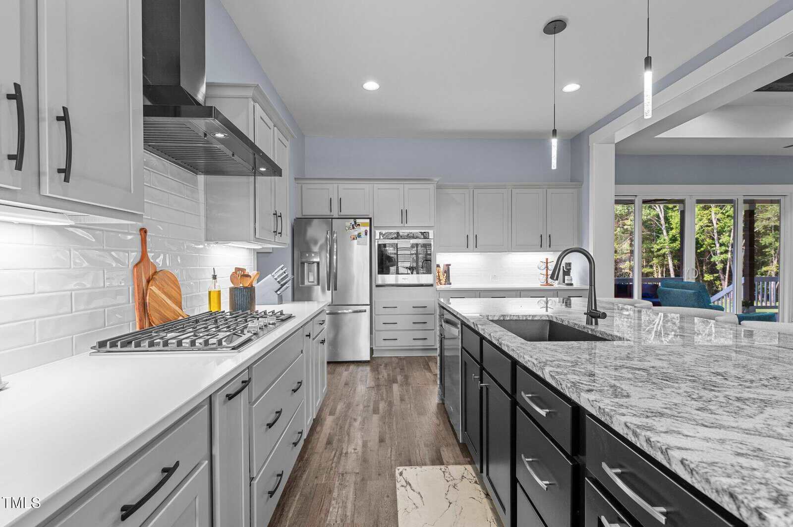 Modern kitchen with light gray cabinets, stainless steel appliances, white tile backsplash, a large center island with a marble countertop, black lower cabinets, and wood flooring. Sliding doors lead to an outdoor patio with greenery.