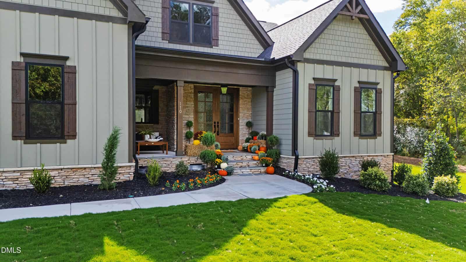 A modern house with gray siding and dark brown trim features a front porch decorated with potted plants and pumpkins. The landscaped yard includes shrubs, flowers, and a green lawn, with a concrete walkway leading to the entrance.