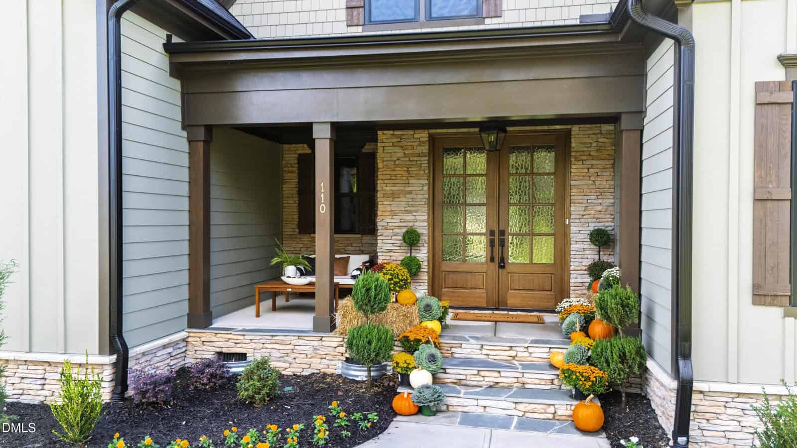 A house front with stone and wood accents, double glass doors, and a covered porch. Steps are decorated with pumpkins, potted plants, and yellow flowers. A bench with pillows sits to the left of the entrance.