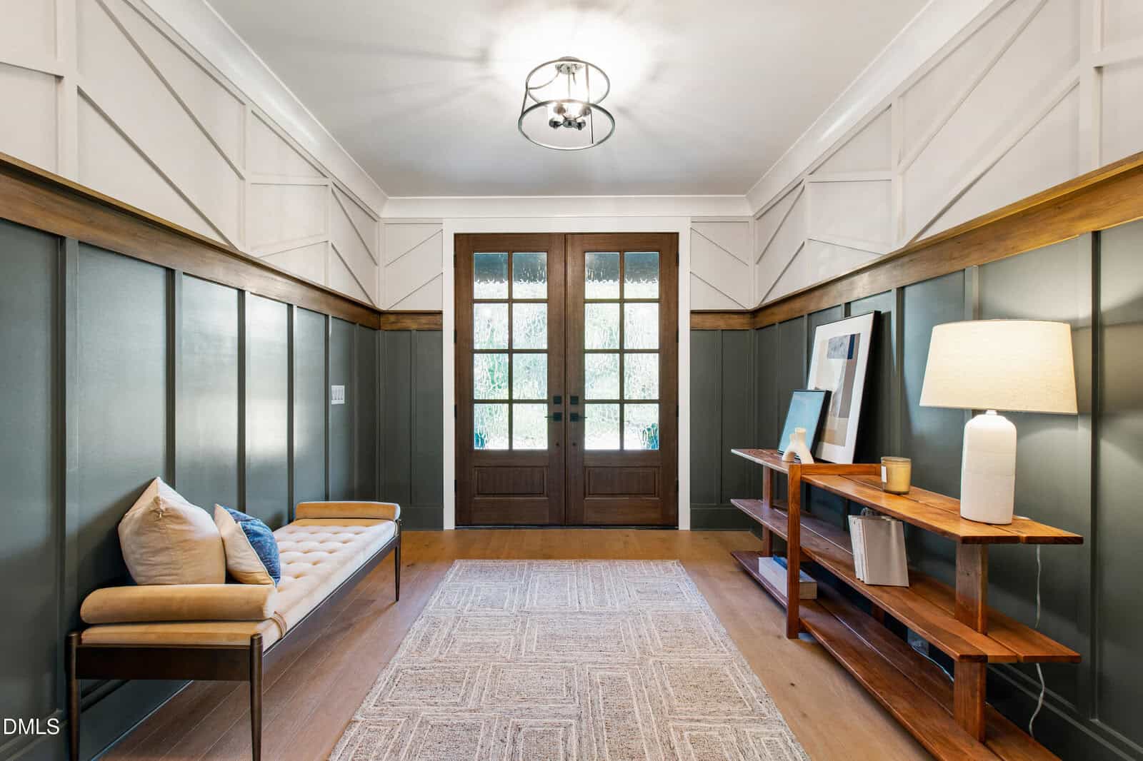 A hallway with double wooden doors, a glass console table with decor and lamp, a light bench with pillows, wood paneling on the walls, a large rug, and a modern ceiling light fixture.