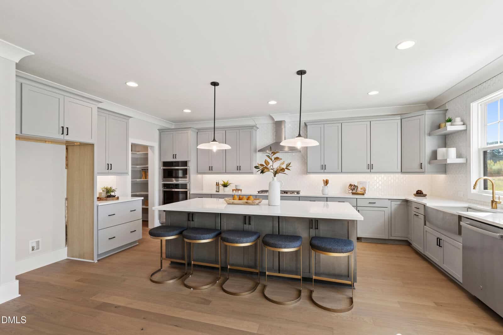 Modern kitchen with gray cabinets, a large island with four blue cushioned stools, pendant lights, stainless steel appliances, white countertops, wood flooring, and a window above a gold faucet sink.
