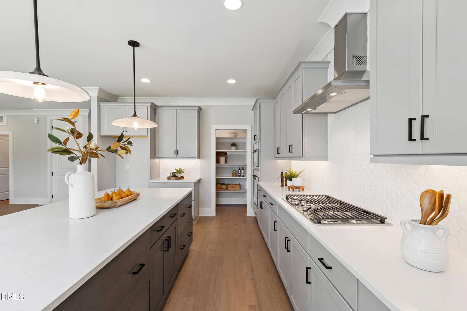 Modern kitchen with gray cabinets, white countertops, wooden floors, a gas cooktop, built-in shelves, and decorative items including a white pitcher with branches and a tray of bread on the island. Bright, natural lighting.