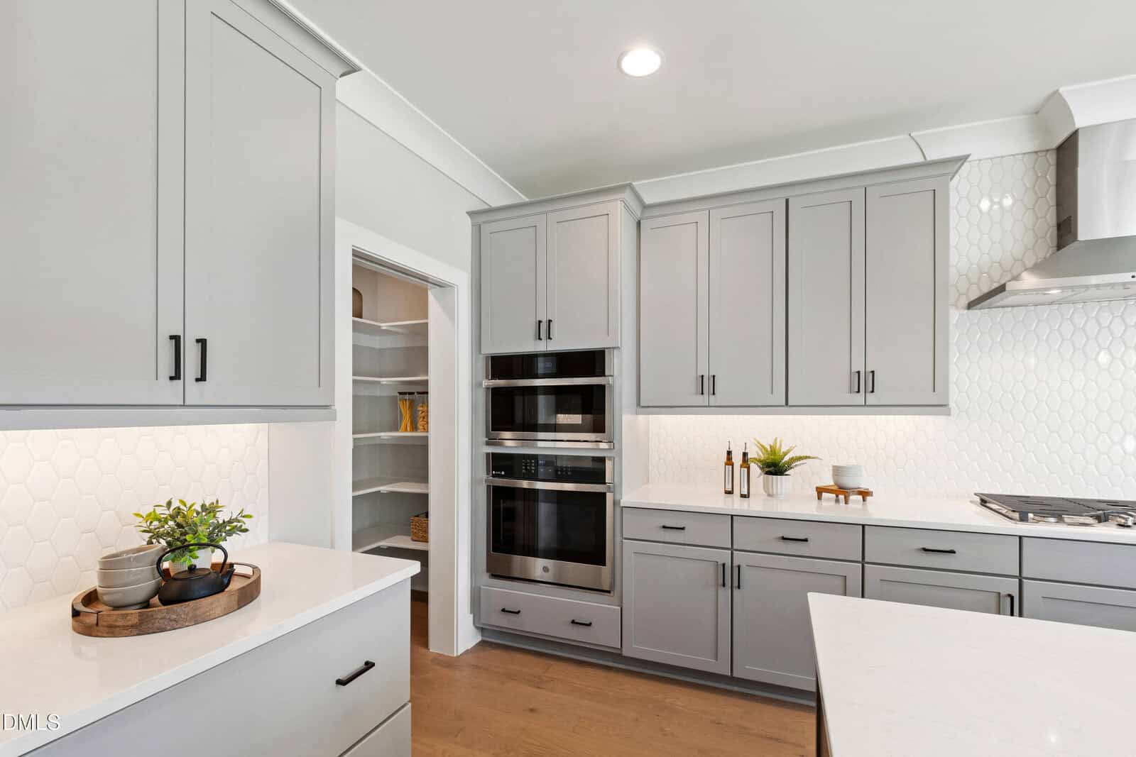 Modern kitchen with gray cabinets, white countertops, built-in double ovens, a visible walk-in pantry, stainless steel gas cooktop, white hexagon tile backsplash, and light wood flooring. Decorative plants are on the counters.