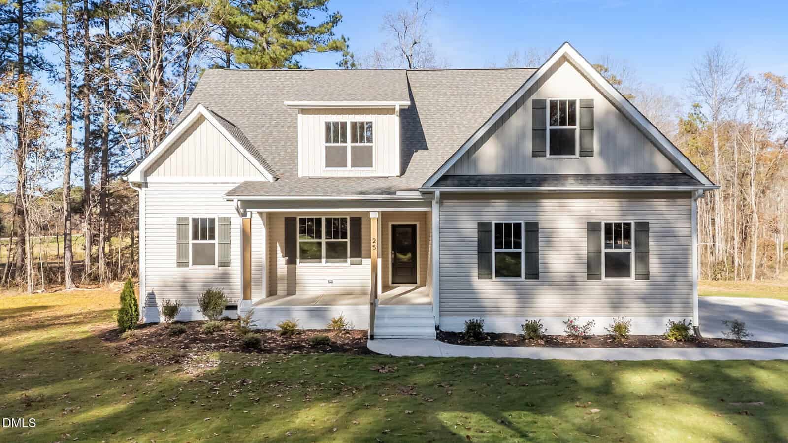 A modern, light gray two-story house with white trim, a covered front porch, dormer windows, and a driveway, surrounded by grass, trees, and a blue sky.