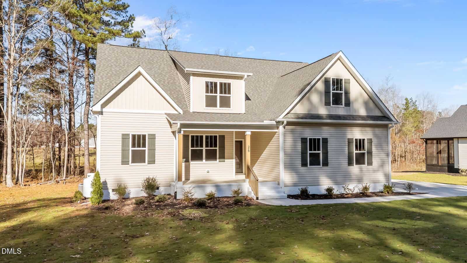 A modern beige two-story house with a covered front porch, white trim, and a gray shingled roof, set on a well-maintained lawn with shrubs and surrounded by tall trees under a blue sky.