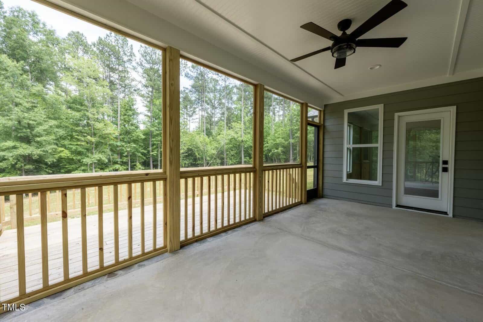 A covered porch with a ceiling fan, screened wooden railing, concrete floor, and view of a wooded backyard. A glass door and large window are set in a green exterior wall.