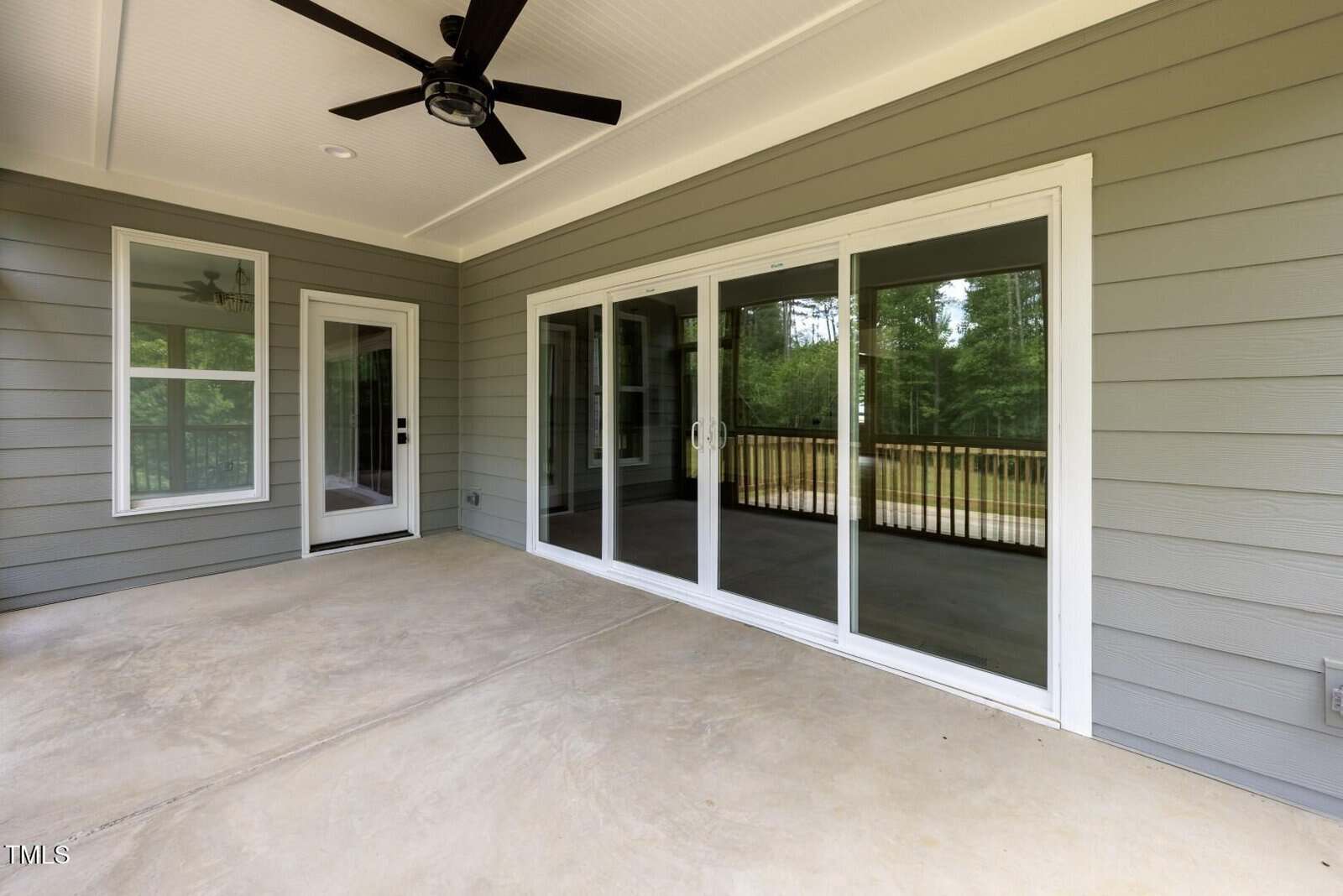 A covered patio features a ceiling fan, gray horizontal siding, a glass door, and large sliding glass doors that open to a deck with a black railing, overlooking a wooded area.
