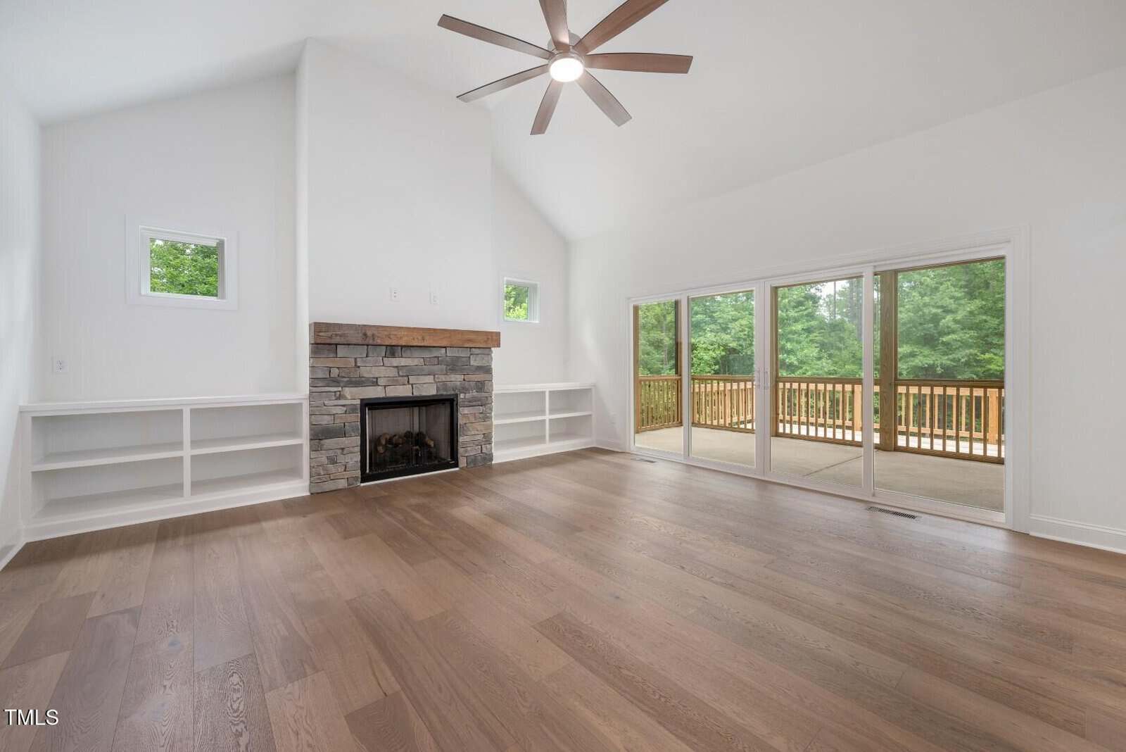 Bright, empty living room with large windows, sliding glass doors to a deck, a stone fireplace, built-in shelves, light wood flooring, and a vaulted ceiling with a modern ceiling fan.