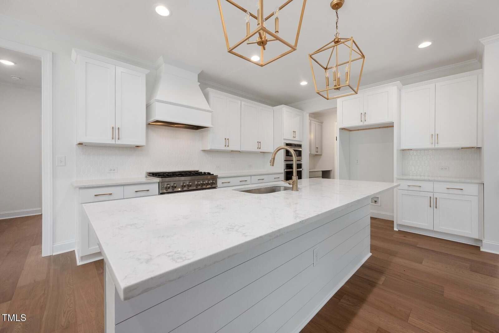 Modern kitchen with white cabinets, a large marble island with a built-in sink, stainless steel appliances, wooden floors, and gold geometric pendant lights hanging from the ceiling.