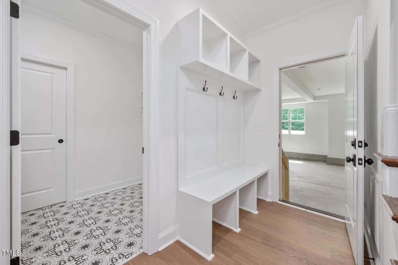 A white mudroom with built-in cubbies, bench, and hooks. To the left is a patterned tile floor room with a white door; to the right is a doorway to a garage with concrete floors and windows.