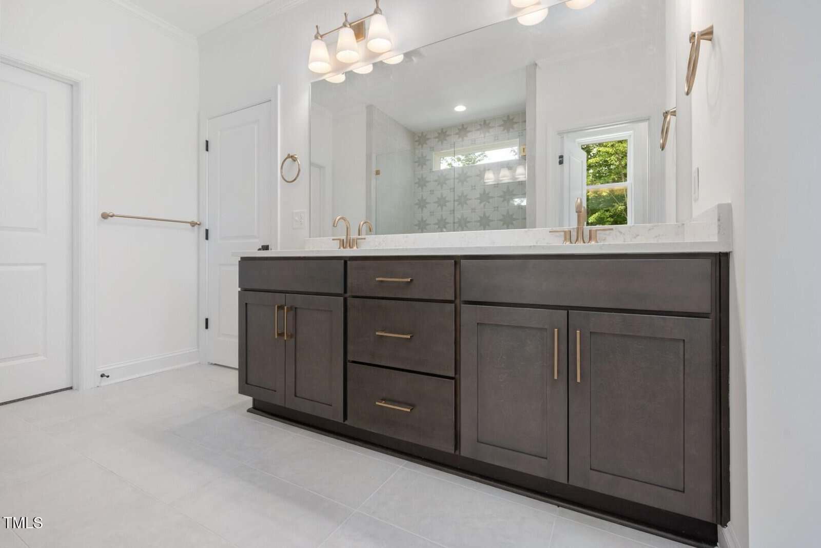 Modern bathroom with a large dark wood vanity, double sinks, gold fixtures, a wide mirror, white walls, and a glass shower with patterned tile reflected in the mirror. A window lets in natural light.